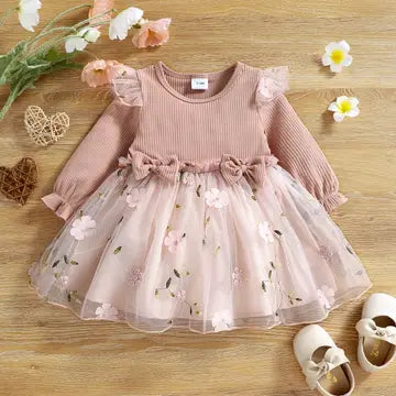Pink children's dress with floral details on a wooden surface with flowers and shoes.