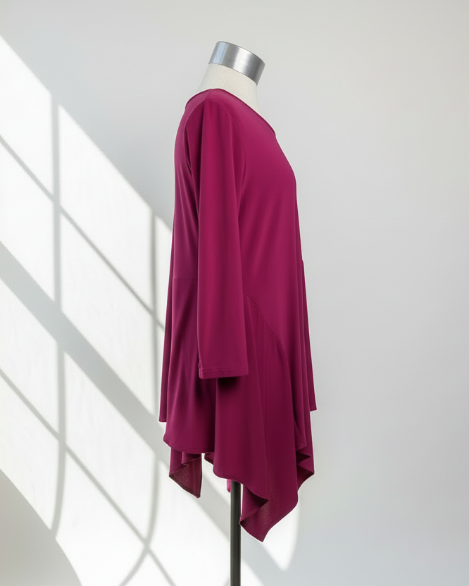 Side profile of vibrant magenta asymmetric tunic on dress form showing flowing drape and handkerchief hem detail, clean white background with natural window light