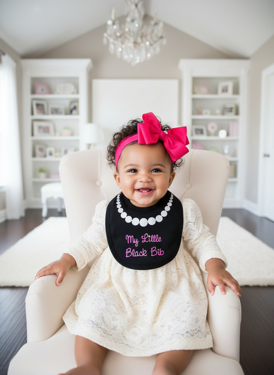 Stylish baby girl with headband wearing black My Little Black Bib with white pearl necklace and hot pink glitter text in upscale boutique setting