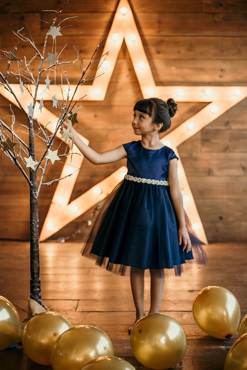 Girl in navy blue capped sleeve dress with rhinestone waist reaching for star ornament