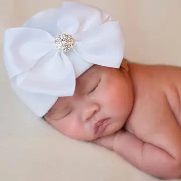 Newborn baby wearing white hospital hat with large rhinestone bow sleeping peacefully