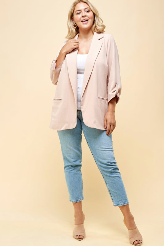 A woman wearing a blush pink blazer with long sleeves, paired with a white top and blue jeans. She is posing with one hand on her hip.