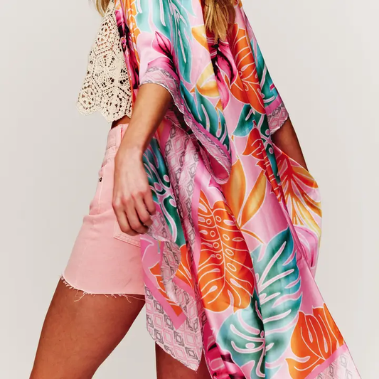This image features a vibrant Tropical Palm Leaves Cover Up Kimono, ideal for adding a bold, exotic touch to beach or resort wear. The kimono displays a lively pattern of tropical palm leaves in various shades of green, orange, and pink, contrasting beautifully against the model's pink shorts.