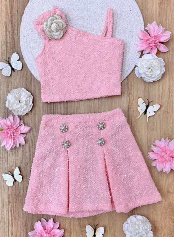 A girls' asymmetric tweed skirt set in pink, with a sleeveless top and a skirt featuring pleats and decorative flowers on the waistband, displayed on a wooden surface surrounded by white and pink flowers and butterflies.