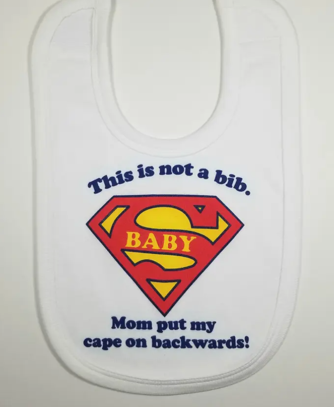 White baby bib with a super hero logo and text 'This is not a bib, BABY. Mom put my cape on backwards!'