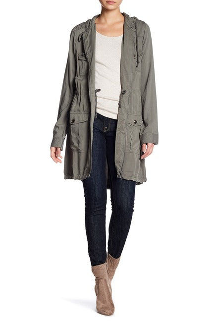Olive green hooded utility jacket worn open over white tee and black jeans, showing relaxed silhouette and multiple cargo-style pockets, styled with tan ankle boots