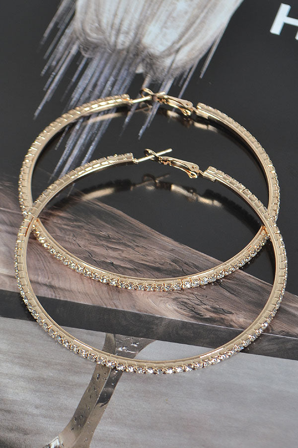 Large gold rhinestone hoop earrings displayed on natural driftwood against dark background, showing 3-inch diameter hoops fully encrusted with clear crystals and secure hinge closure