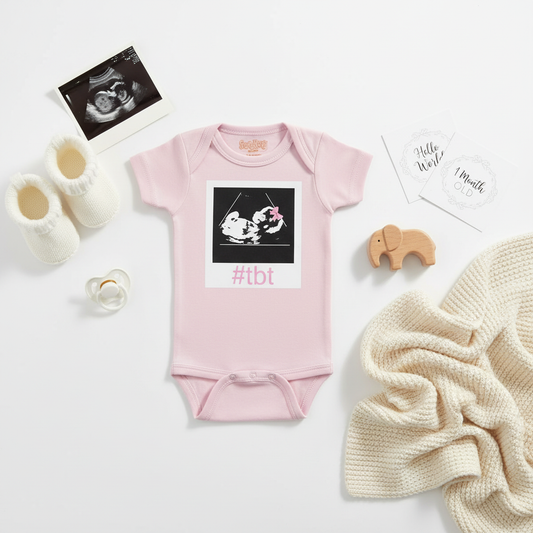 Pink baby onesie with sonogram ultrasound image and #TBT text, styled with baby booties, pacifier, and soft blanket on white background