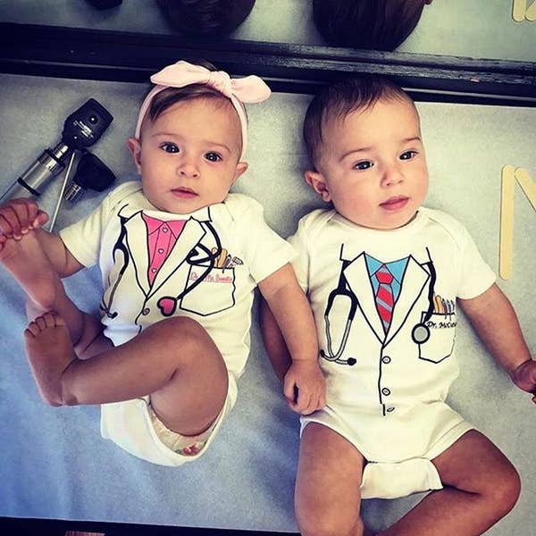 Two babies wearing white onesies with a doctor's costume design, including a stethoscope and name tag, with one baby wearing a pink bow headband.