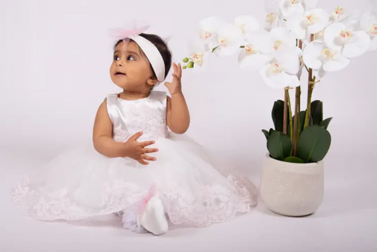 Baby wearing white lace christening dress with pink accents and matching headband, sitting next to white orchids