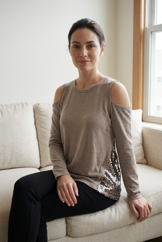 Taupe cold-shoulder top with metallic sequin side panels on a model seated naturally on a cream couch