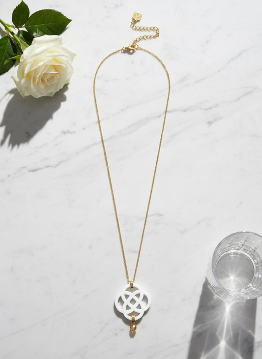 Gold chain necklace with white acrylic quatrefoil pendant and crystal accent styled with white roses and crystal glass on marble surface