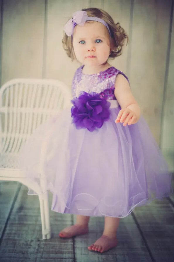 A baby wearing a purple party dress with sequin detailing and a large flower on the waist, standing next to a white chair.