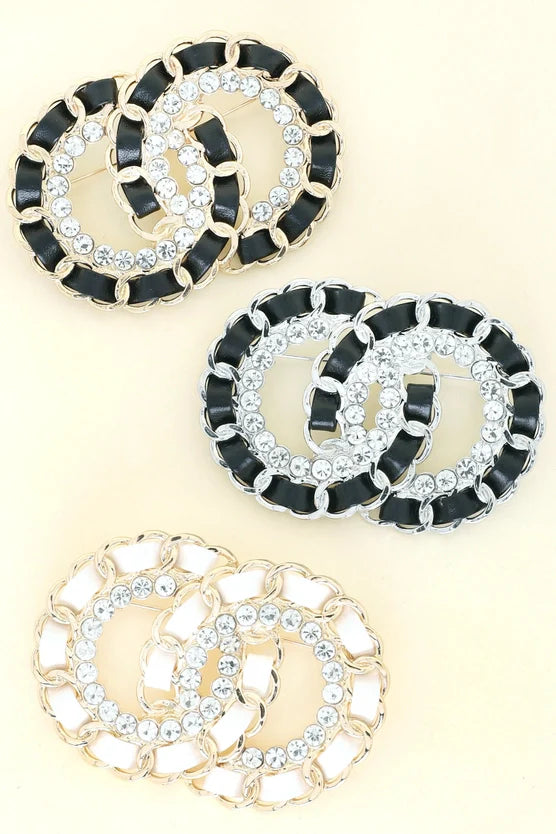 Three designer brooches with interlocking CC logo design in black and white enamel with crystal rhinestone embellishments on cream background