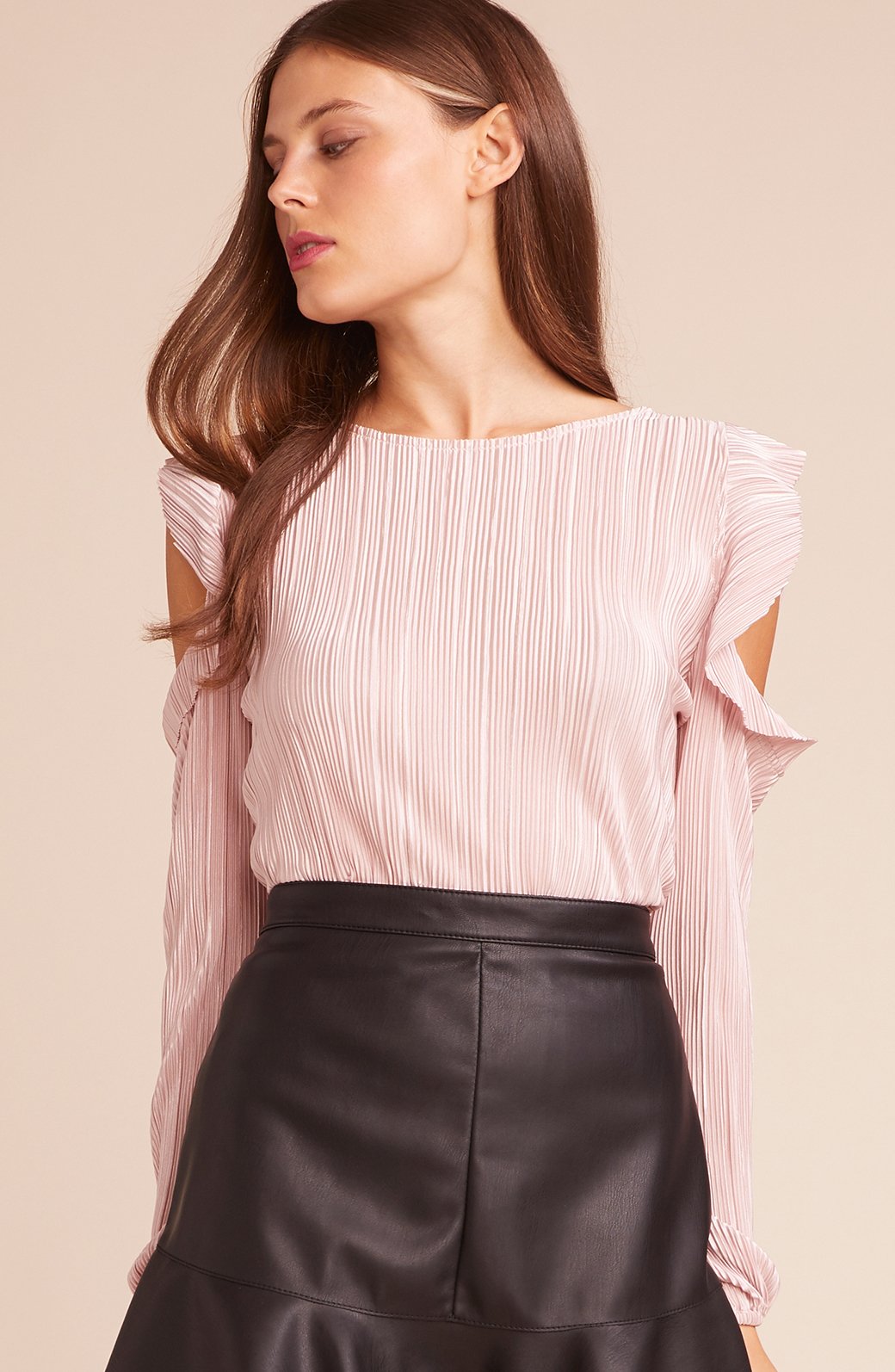 A woman wearing a light pink pleated long-sleeve top with ruffled shoulder details and a black faux-leather skirt, set against a solid beige background.