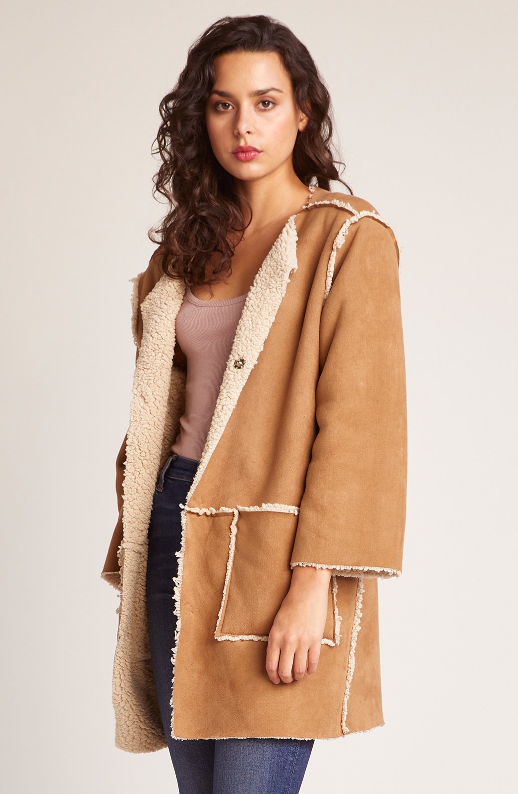 Woman wearing camel reversible sherpa coat with faux suede exterior, cream fleece trim, hooded collar and patch pockets styled with dark jeans