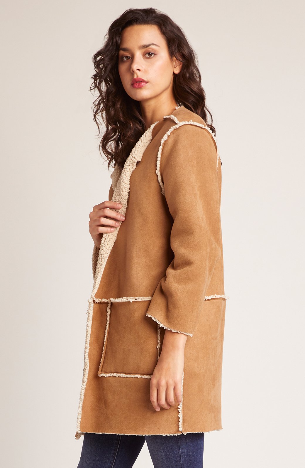 Side view of woman wearing camel reversible sherpa coat showing relaxed fit, raw frayed edges and mid-thigh length