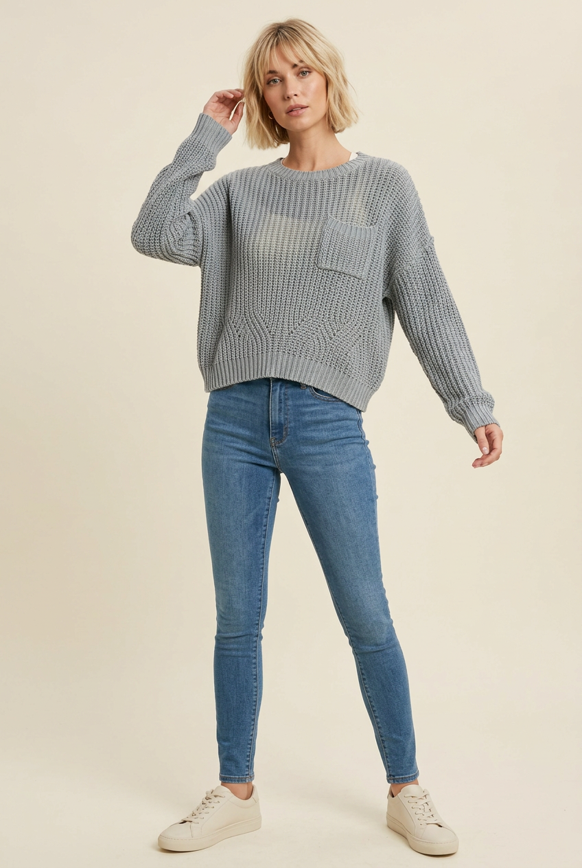 Dusty blue open-knit pocket sweater with a relaxed near-cropped silhouette, styled on a blonde model with skinny jeans and ivory sneakers.