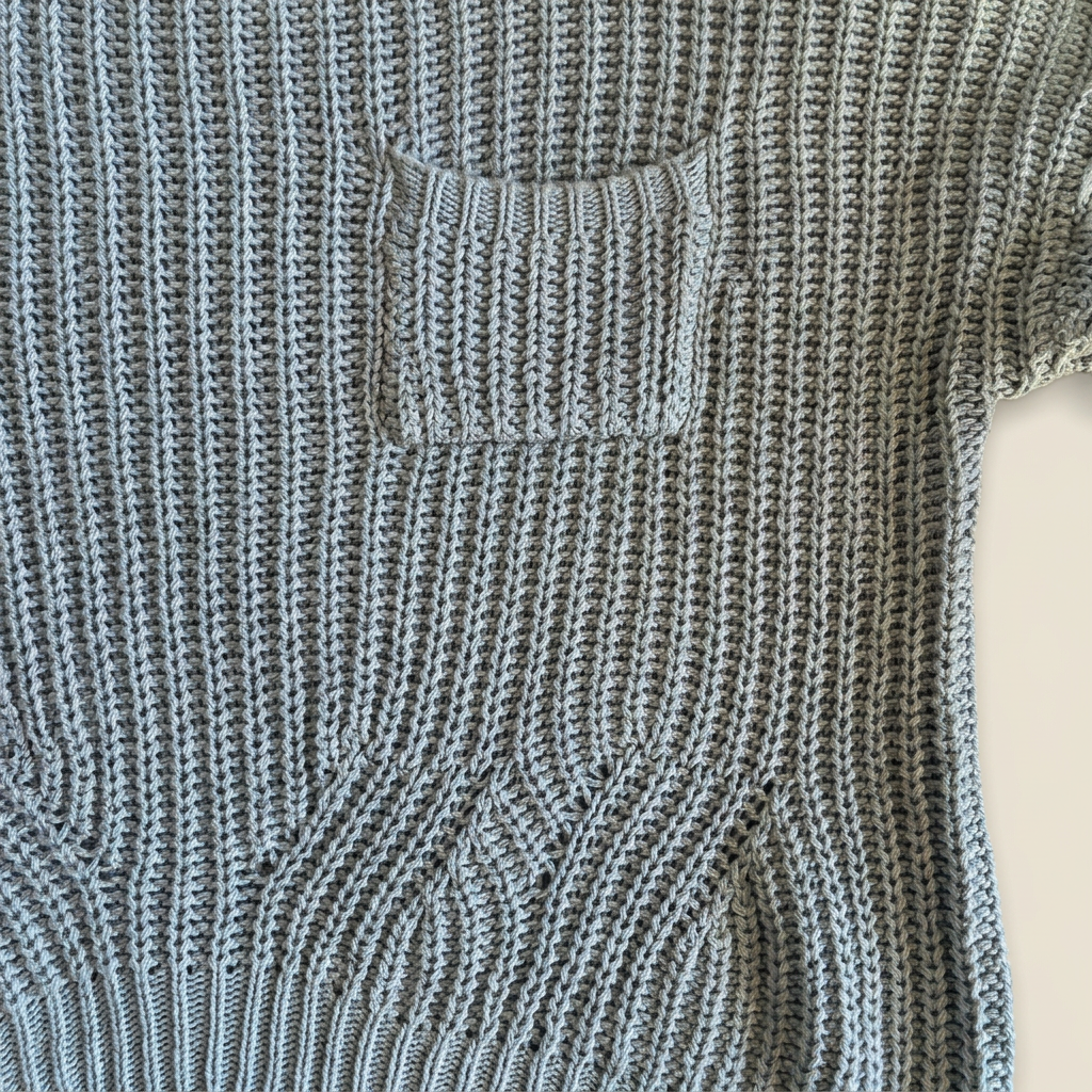 Close-up of dusty blue open-knit sweater showing the chest patch pocket, ribbed pocket edge and curved stitch detailing above the ribbed hem.