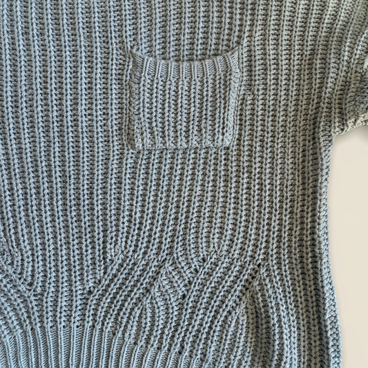 Close-up of dusty blue open-knit sweater showing the chest patch pocket, ribbed pocket edge and curved stitch detailing above the ribbed hem.