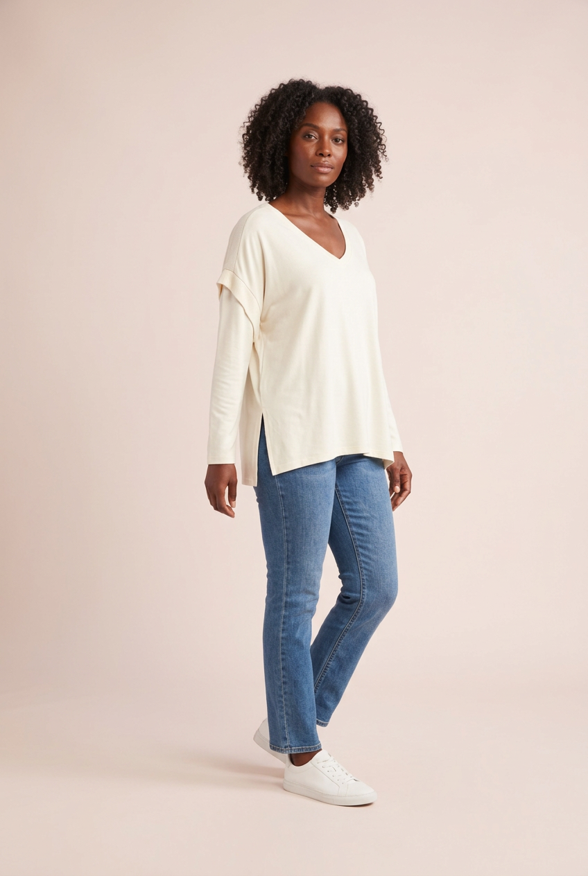 Ivory relaxed V-neck long sleeve top with side slits, styled with blue jeans and white sneakers, three-quarter model view on a pink backdrop.