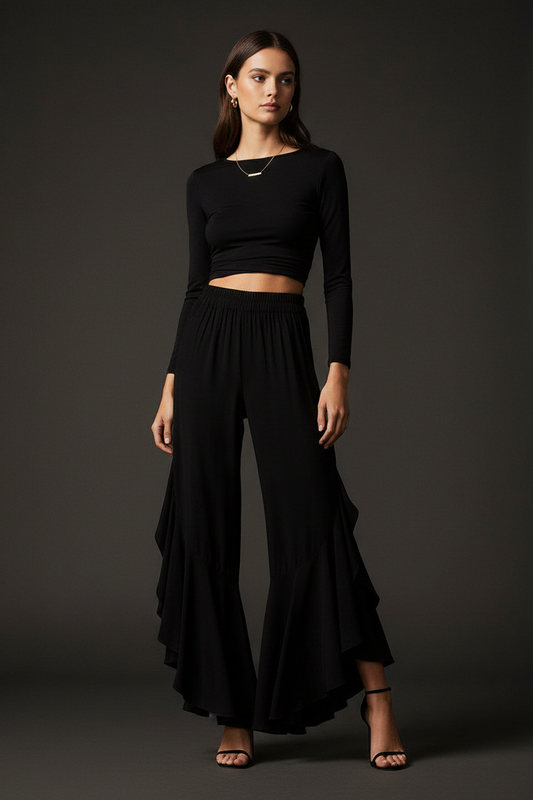 Woman wearing black ruffle hem wide-leg flowy pants with sleek crop top and strappy heeled sandals in moody studio setting – evening trouser by Pinktini