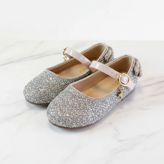 Girls' silver and blush rhinestone Mary Jane dress shoes on white marble surface – professional product shot showing bow back and gold ankle strap – by Pinktini
