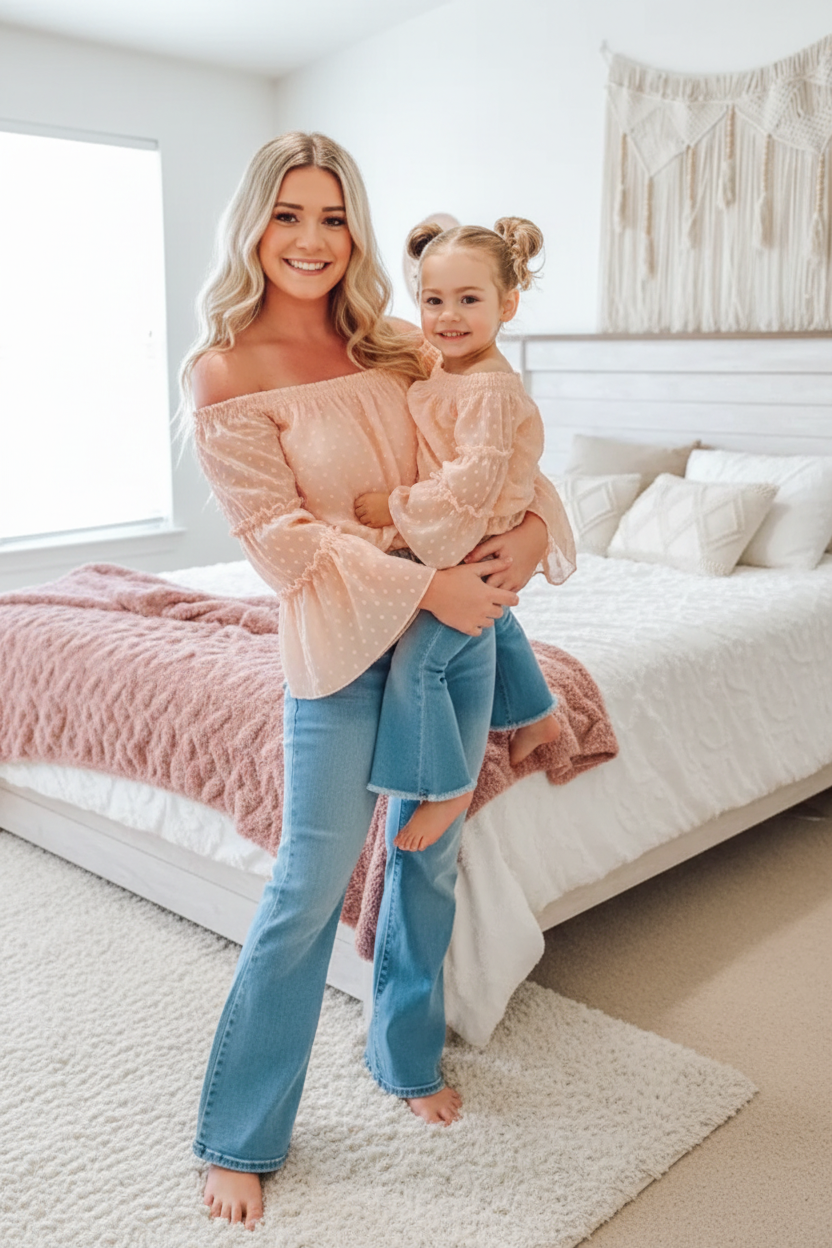 Mother and daughter wearing matching peach off-the-shoulder Mommy and Me tops with blue jeans, sitting together on a bed
