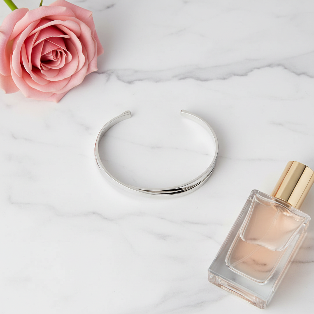 Silver crossed flat metal bangle bracelet on white marble surface with pink rose and perfume bottle