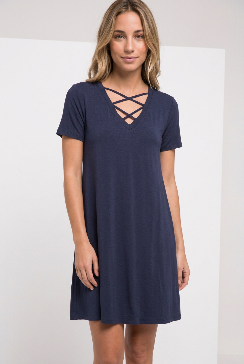 Navy short-sleeve T-shirt swing dress with double-crisscross V-neck straps, worn by a blonde model in a relaxed upright pose against a pale studio backdrop.