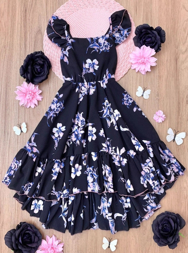 A navy blue hi-lo ruffle dress with floral patterns, displayed on a wooden surface with pink and black decorative flowers around it.