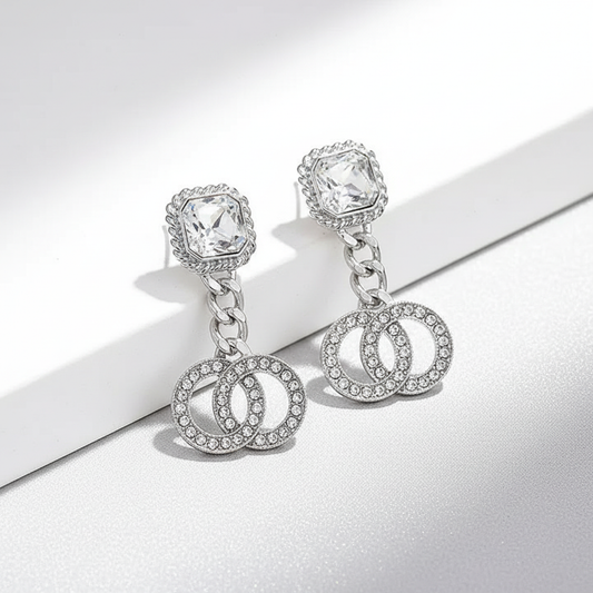 Pair of silver double circle drop earrings on bright white background, featuring square crystal stud tops with rope frames, chain connectors, and 1.25-inch wide rhinestone interlocking circles