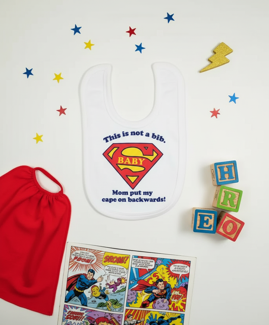 White Super Baby bib with superhero shield surrounded by red cape, comic book, and playful props on white background