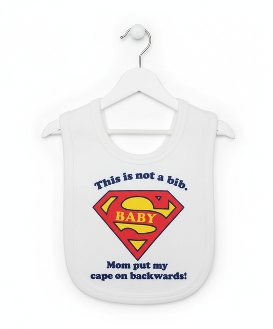White baby bib with red and yellow Superman BABY shield and 'This is not a bib Mom put my cape on backwards' text hanging on white hanger
