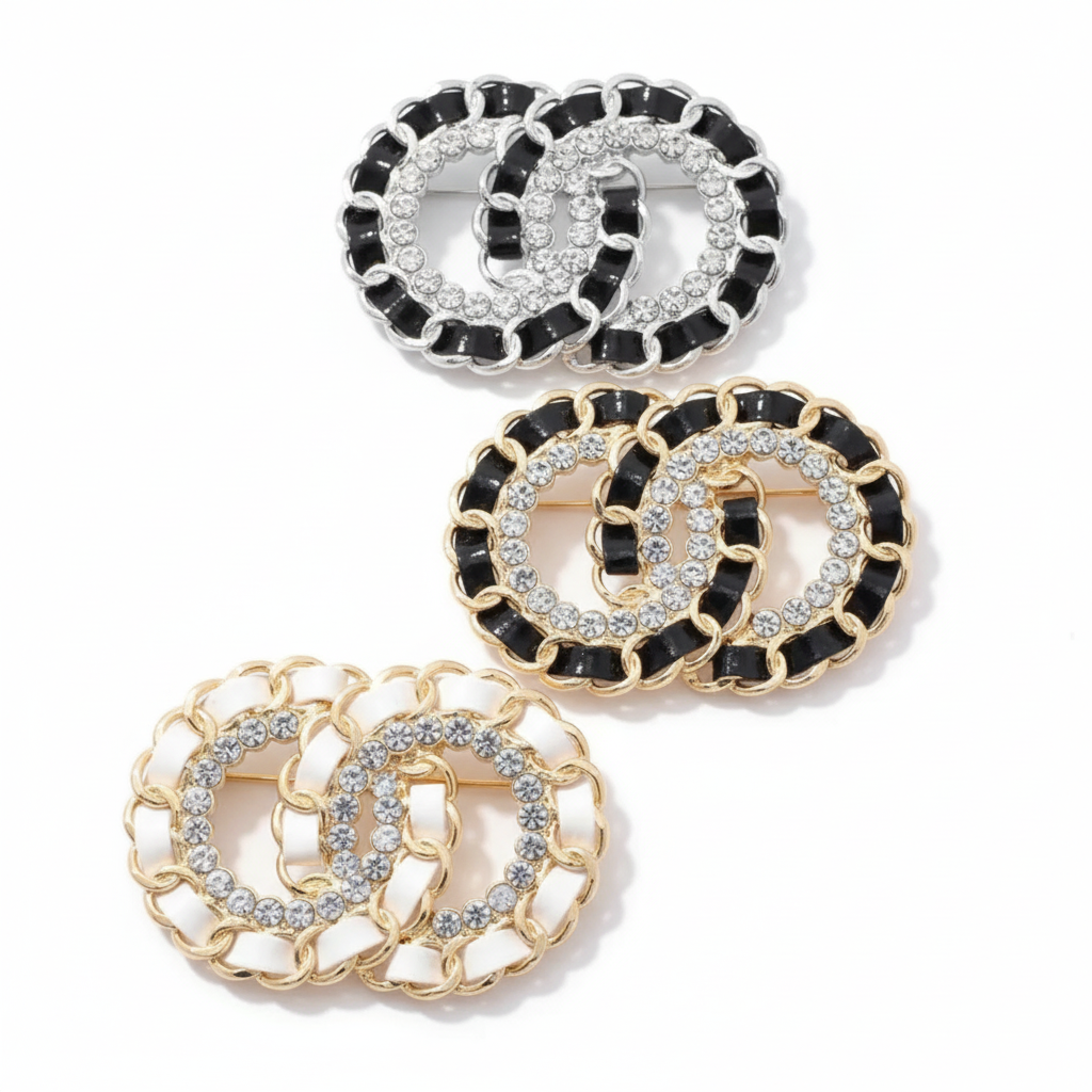 Three interlocking CC logo chain-link brooches showing color variations - black enamel with silver chain, black enamel with gold chain, and white enamel with gold chain, all with clear rhinestone CC logos on white background