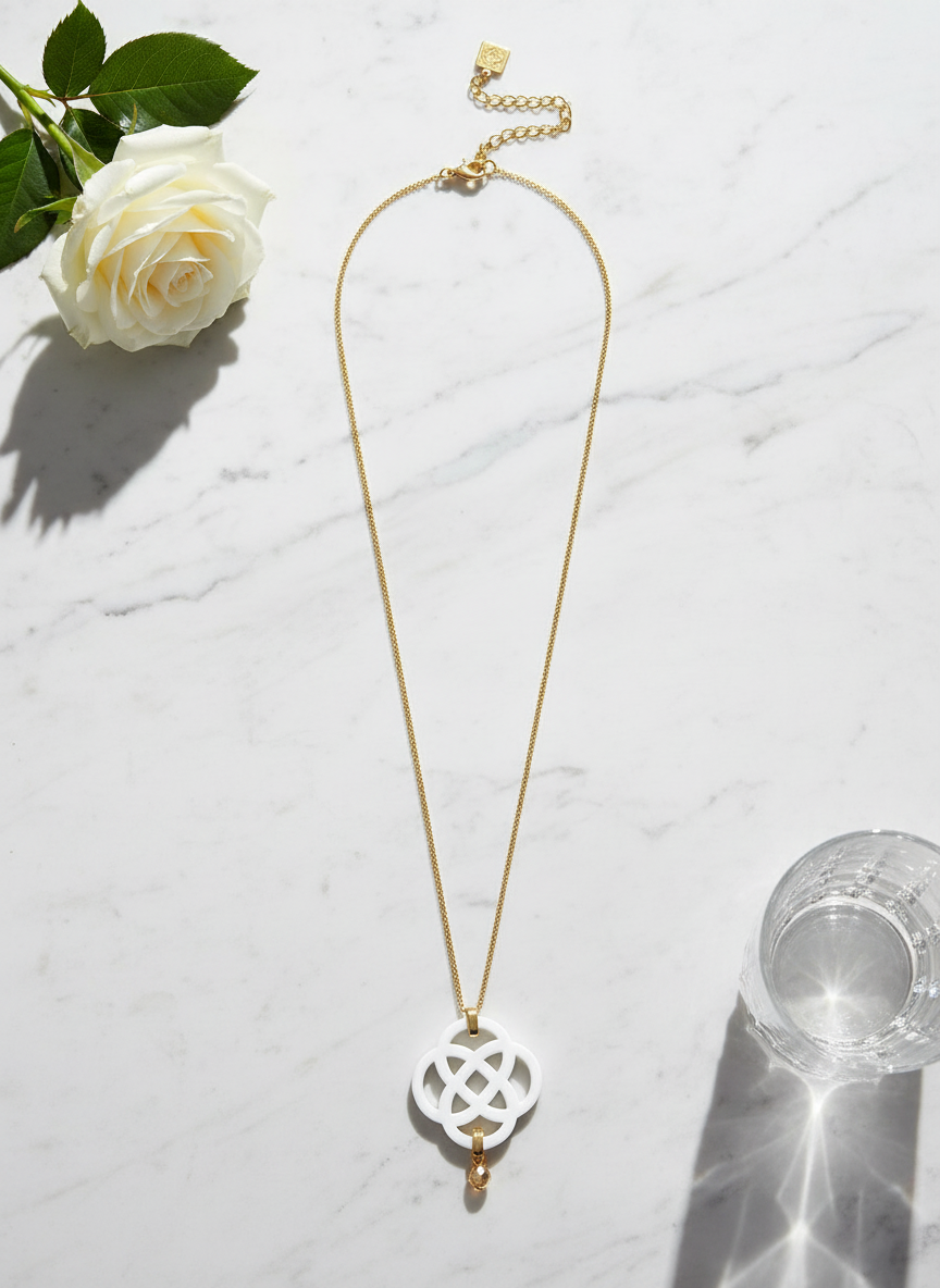 Gold chain necklace with white acrylic quatrefoil pendant and crystal accent styled with white roses and crystal glass on marble surface