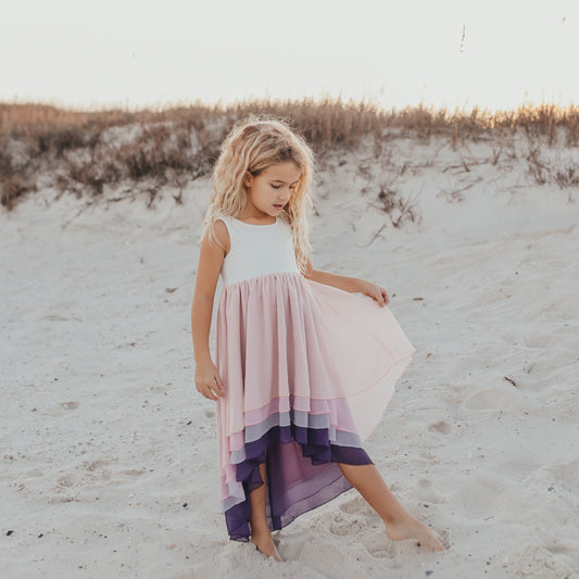 A girl standing on sand at the beach wearing a hi-lo tiered lavender ombre dress with a white bodice and sleeveless design.